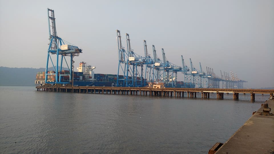 Blue ship-to-shore cranes over a container ship berthed along a long pier at Jawaharlal Nehru Port, Navi Mumbai, in morning haze.